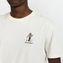Load image into Gallery viewer, A close-up of a man wearing a cream-colored Revolution t-shirt with a small graphic on the left chest depicting a worker and the brand name.