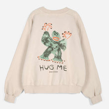 Load image into Gallery viewer, A beige crewneck sweatshirt shown from the back, featuring a large graphic of a green monster with orange accents and the text 'HUG ME' printed below.