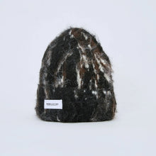 Load image into Gallery viewer, A fuzzy knit beanie hat with a mottled black, brown, and white pattern, featuring a small white rectangular brand label near the bottom left, set against a plain light gray background.